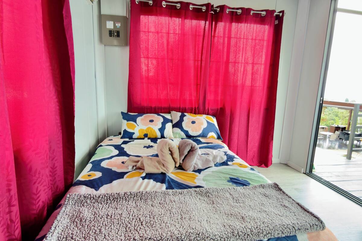 A bed with a colorful floral bedspread, red curtains, and a view of the outdoors.