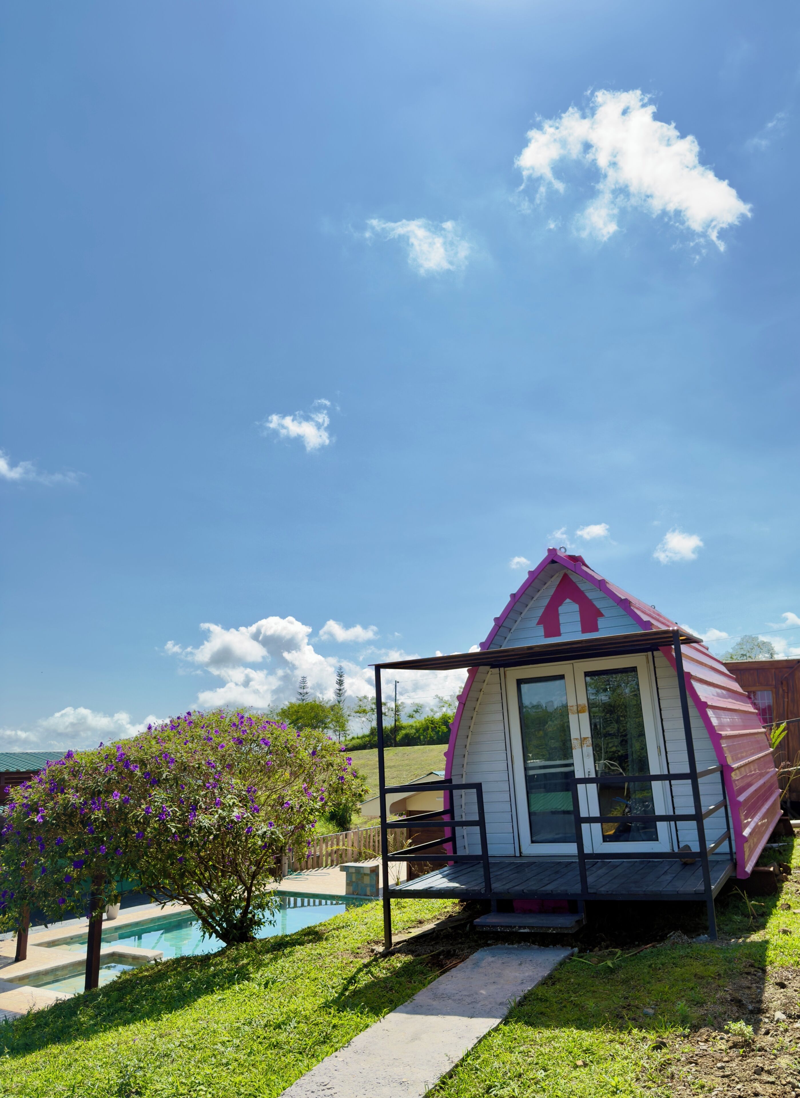 A small, single-story house with a pink roof, a visible front door, and a pool in the background.