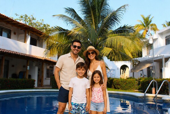 A family by a pool with a white building, palm trees, and a tiled roof. - Casa Coral (Manzanillo)