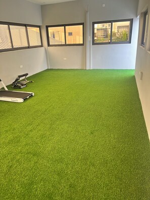 Ein Zimmer mit künstlichem Grünteppich, Laufband und Fitnessgeräten.