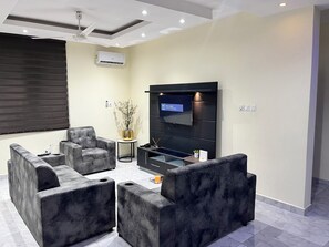 A modern living room with a flat-screen TV, grey upholstered sofas, a small side table, and a potted plant.