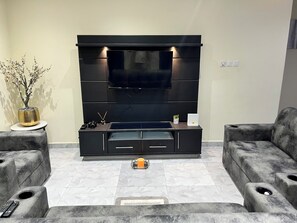 Ein modernes Wohnzimmer mit Flachbildfernseher, dunklem Holz-Entertainment-System, zwei grauen Sofas, einem kleinen Tisch mit einer Vase und einer Topfpflanze.
