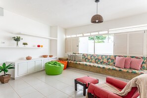 Ein helles Wohnzimmer mit einer Couch, einem grünen Sitzsack, einem roten Sessel und einem kleinen Tisch. Es gibt Blumentöpfe und ein Fenster mit Fensterläden.