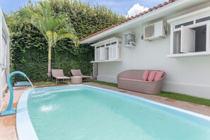 Ein Schwimmbecken mit klarem, blauem Wasser, umgeben von einem weißen Gebäude mit einem Ziegeldach, einem Korbsessel mit gestreiften Kissen und zwei Liegestühlen.