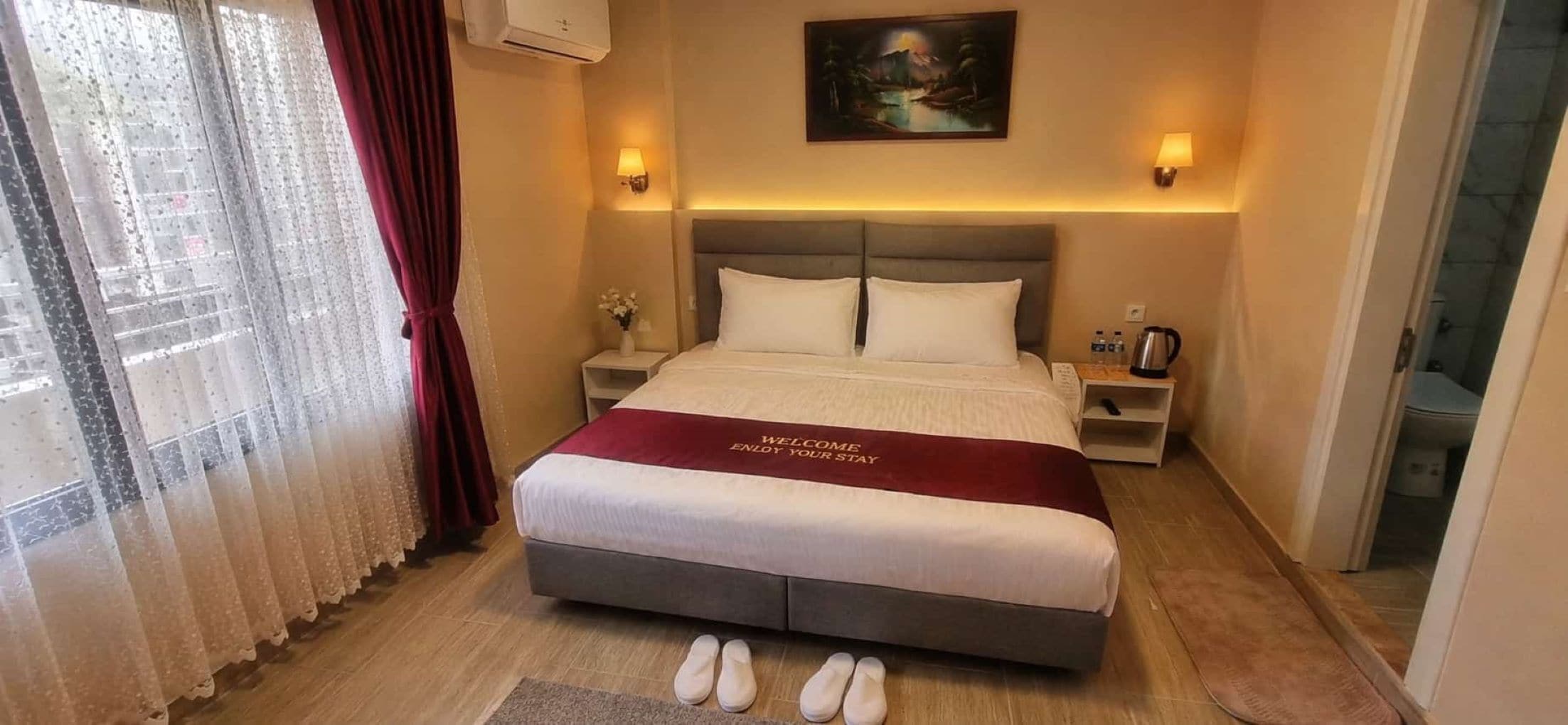 Habitación de hotel con una cama grande, una mesita de noche con lámpara, un cuadro en la pared y vistas al exterior a través de una ventana con cortinas.