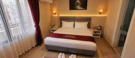 Habitación de hotel con una cama grande, una mesita de noche con lámpara, un cuadro en la pared y vistas al exterior a través de una ventana con cortinas.