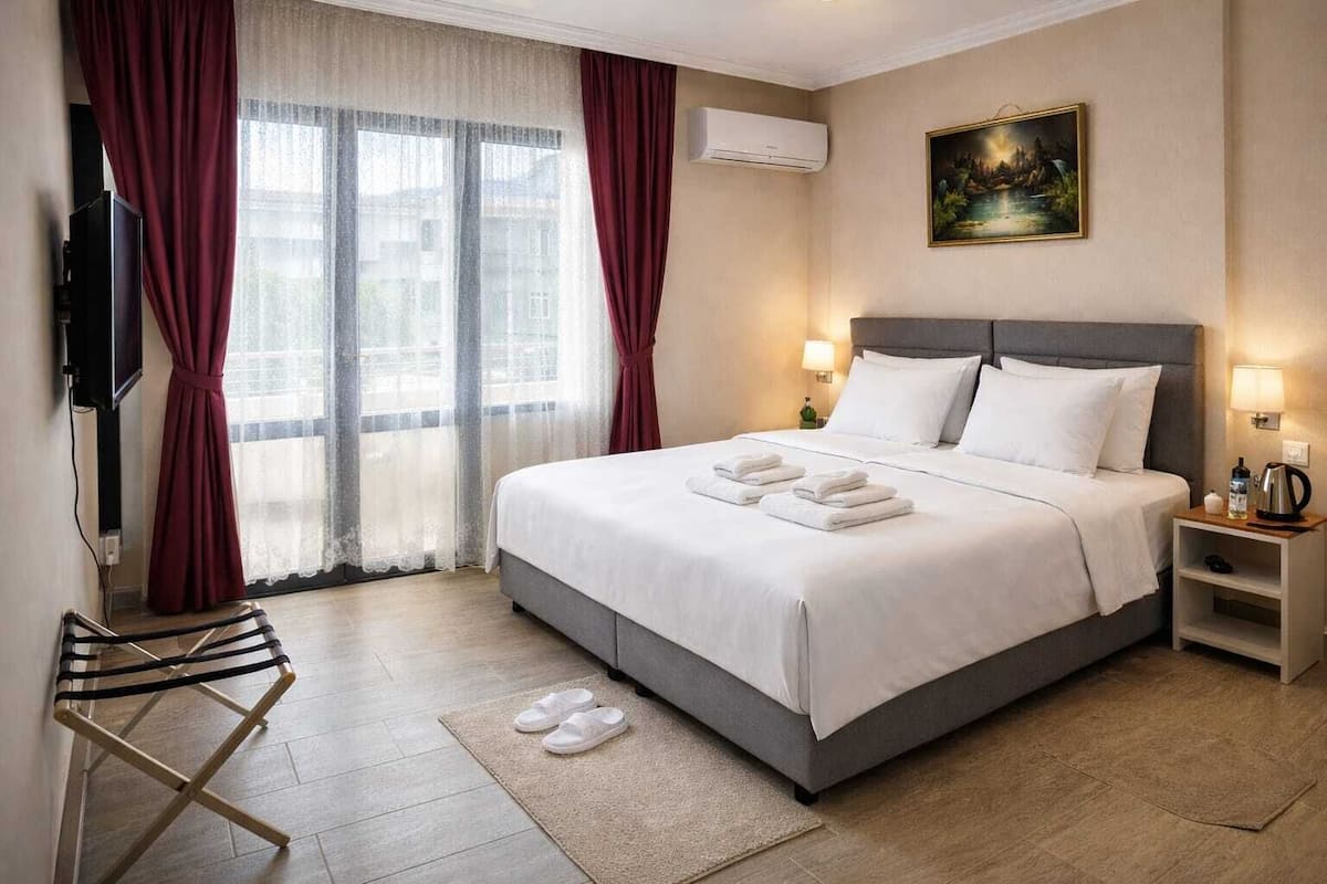 A hotel room with a bed, a TV, a painting, and a view of buildings through the window.