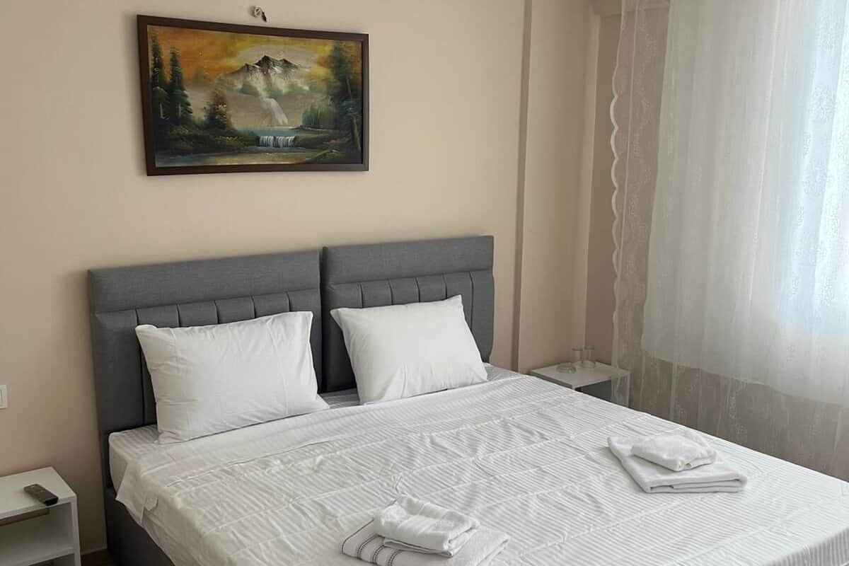 A neatly made bed with white linens, a grey headboard, and a painting of a mountain landscape on the wall.