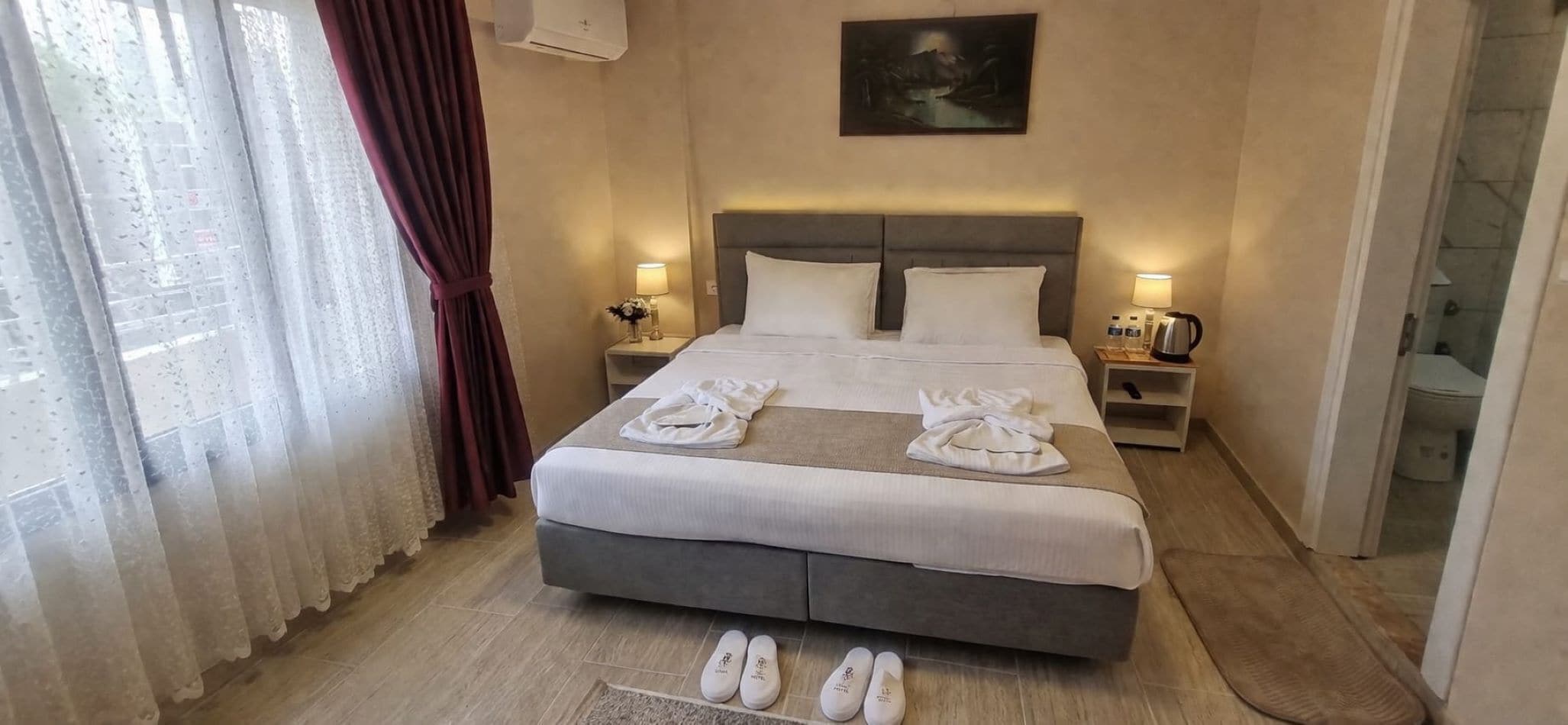 Una habitación de hotel con una cama, dos mesitas de noche, un aparador, un cuadro en la pared y vistas a través de una ventana con cortinas.