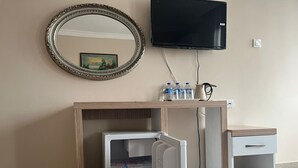 Una habitación con escritorio de madera, espejo, televisor, un refrigerador pequeño y un mueble bajo.