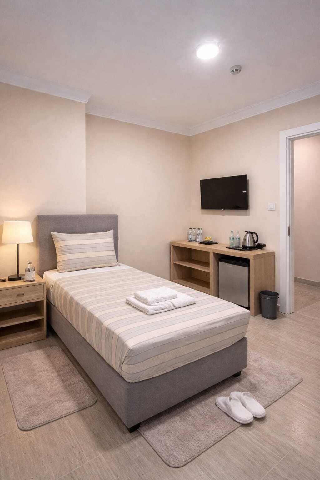 Habitación de hotel ordenada con cama, mesita de noche, lámpara, televisor, escritorio y un pequeño cubo de basura.