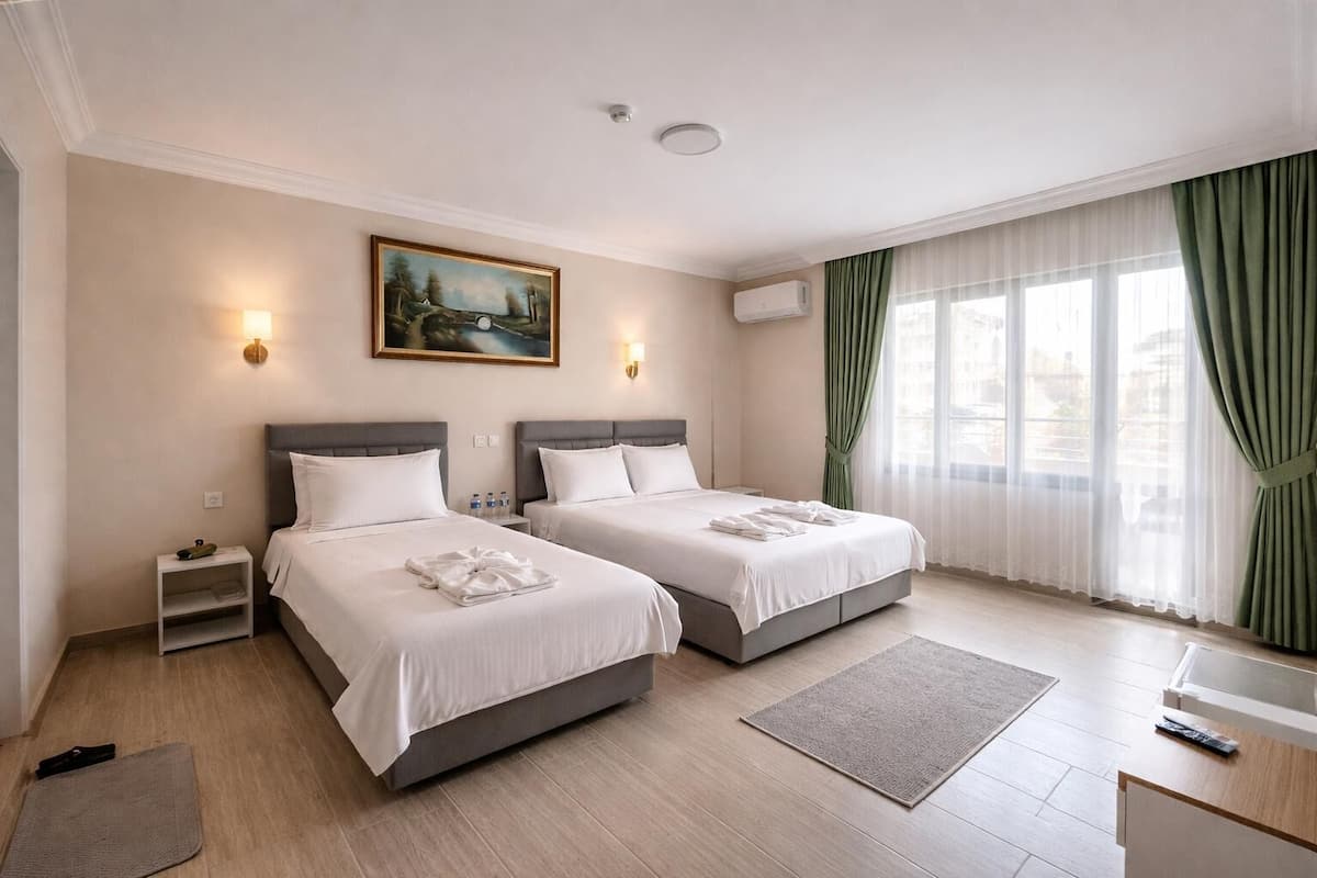 A hotel room with two beds, a painting on the wall, an air conditioning unit, a window with green curtains, and a small bedside table with a lamp.