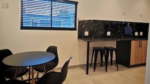 A modern kitchen with a black countertop, a window with blinds, and a dining area with a round table and chairs. - Room-king Deluxe Apartment-style Room (Cape Town)