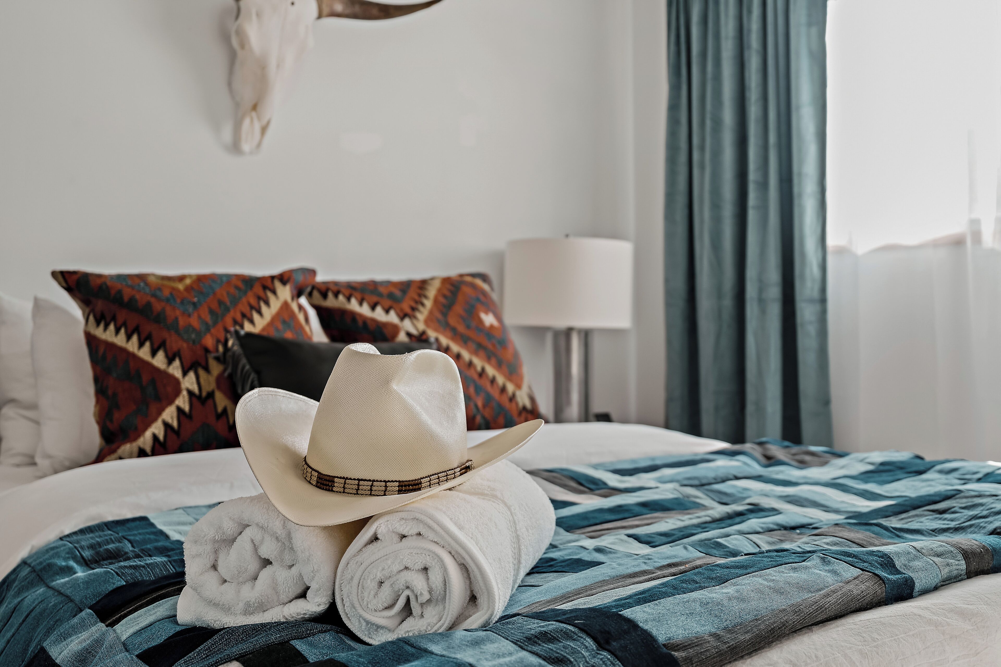 A neatly made bed with a patterned blanket, a rolled white towel, and a cowboy hat.