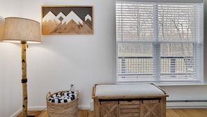 Una stanza con una lampada, un quadro appeso al muro, una finestra con persiane, un cesto di vimini e una panca di legno.