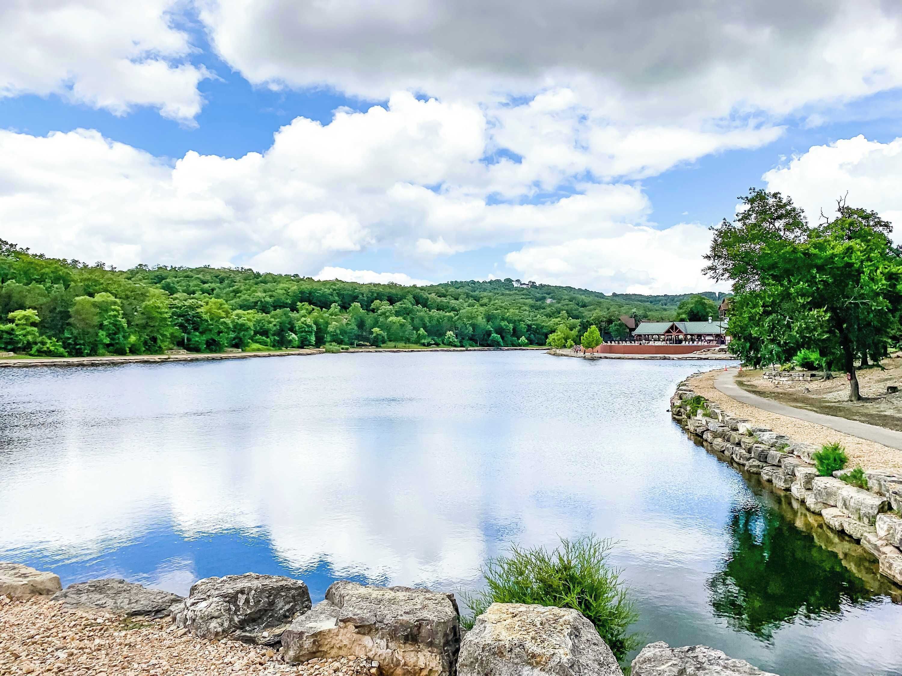 A serene lakeside view with a stone pathway, lush greenery, and a distant building.