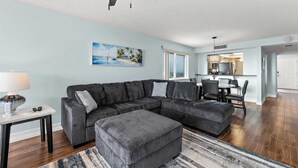 A living room with a sectional sofa, ottoman, and a dining area with chairs and a table. - Oceanview I 3rd FL I Quiet Beach Escape I Beach (Cocoa Beach)