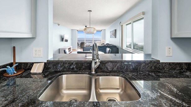 A modern kitchen with a double sink, granite countertops, and a view to the living area. - Oceanview I 3rd FL I Quiet Beach Escape I Beach (Cocoa Beach)