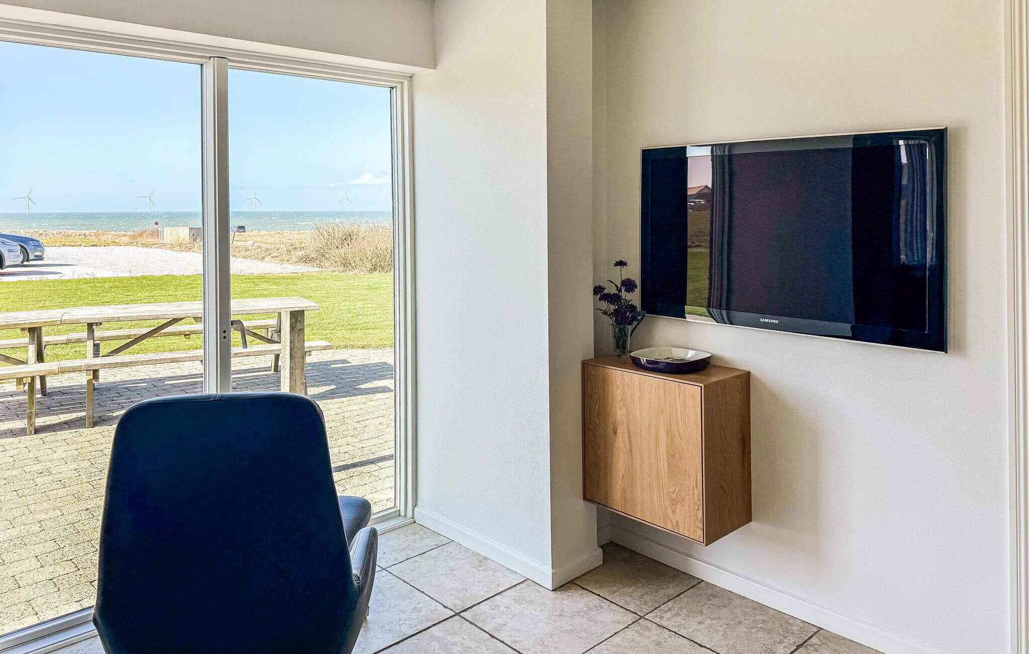 Een kamer met een groot raam, een flatscreen televisie aan de muur, een houten kastje onder de televisie, en een stoel voor het raam.