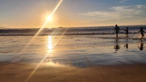 A beach scene with people playing near the water, a clear sky, and the sun reflecting on the wet sand. - Apartment close to the beach, restaurant and beautiful sunsets. (Viña del Mar)