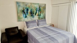 A bedroom with a bed, two pillows, a chair, a painting on the wall, and a window with blinds. - Apartment close to the beach, restaurant and beautiful sunsets. (Viña del Mar)