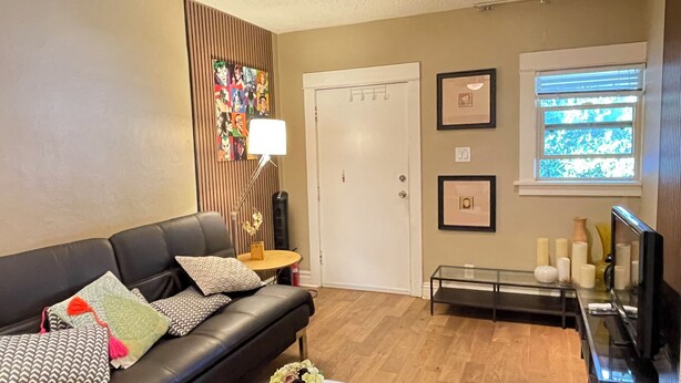 A living room with a black leather sofa, a round coffee table with a vase, a lamp, framed pictures on the wall, and a window with blinds.