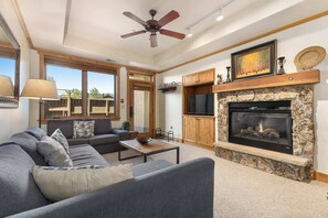 A living room with a fireplace, a ceiling fan, and a large window. - Aspen Oasis | 2BR, Private Hot Tub, Resort Pools (Steamboat Springs)