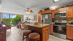 A kitchen with wooden cabinets, a stainless steel refrigerator, a microwave, and an oven. There is a bar area with stools and a ceiling fan. - Double Master 2BR Villa | Pacific Shores | Kihei (Kihei)