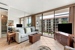 A living room with a sofa, coffee table, TV, and a balcony with a view of a building. - Top Floor Ocean View | Central AC | Walk to Beach (Kihei)