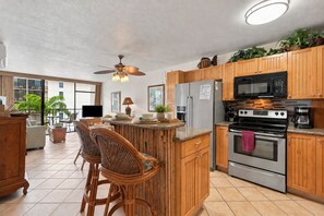 A kitchen with wooden cabinets, a stainless steel refrigerator, a microwave, and an oven. There is a bar area with wicker chairs and a ceiling fan. - Top Floor Ocean View | Central AC | Walk to Beach (Kihei)