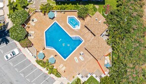 An aerial view of a swimming pool with a geometric shape, surrounded by lounge chairs and umbrellas, with a parking area to the left. - Top Floor Ocean View | Central AC | Walk to Beach (Kihei)