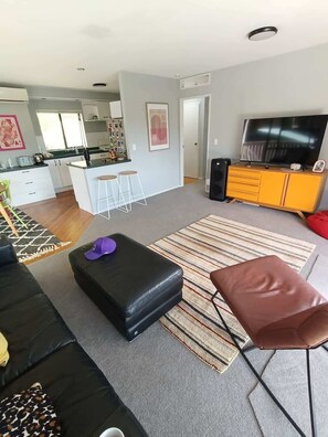 A modern living room with a sofa, ottoman, and a TV on a wooden cabinet. - Hosts on the Coast Pohutukawa Perch (Whitianga)