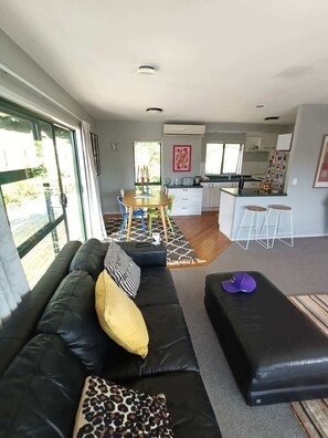 A modern living room with a black leather sofa, a leopard print pillow, and a black ottoman. There is a dining area with a wooden table and colorful chairs. A kitchen with white cabinets and a bar area is visible in the background. - Hosts on the Coast Pohutukawa Perch (Whitianga)