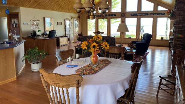 A dining area with a round table, chairs, a vase with sunflowers, and a view of the beach through large windows. - Enjoy a bed & breakfast with view of Lake Huron, sunrises and eagles! (Ocqueoc)