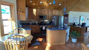 A kitchen with wooden cabinets, a central island, a dining area with a table and chairs, and a potted plant. - Enjoy a bed & breakfast with view of Lake Huron, sunrises and eagles! (Ocqueoc)