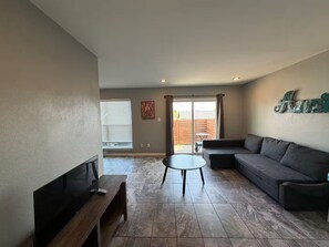 Ein Wohnzimmer mit einem Ecksofa, einem kleinen runden Tisch, einem Flachbildfernseher und einer Glasschiebetür, die zu einem Außenbereich führt.