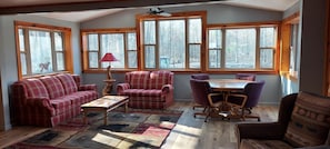 Una habitación acogedora con un conjunto de sofá, una mesa y sillas, ventanas amplias con vistas a árboles y un ventilador de techo.