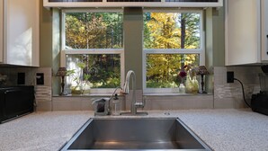 A kitchen with a stainless steel sink, two windows with a view of autumn trees, and a vase with flowers on the windowsill.