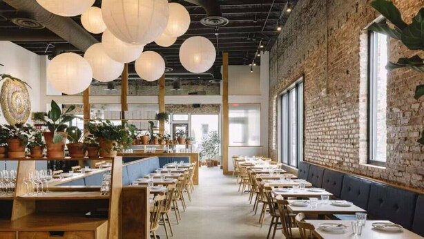 Een restaurant met lange tafels, houten stoelen, hangende papieren lampions en potplanten.