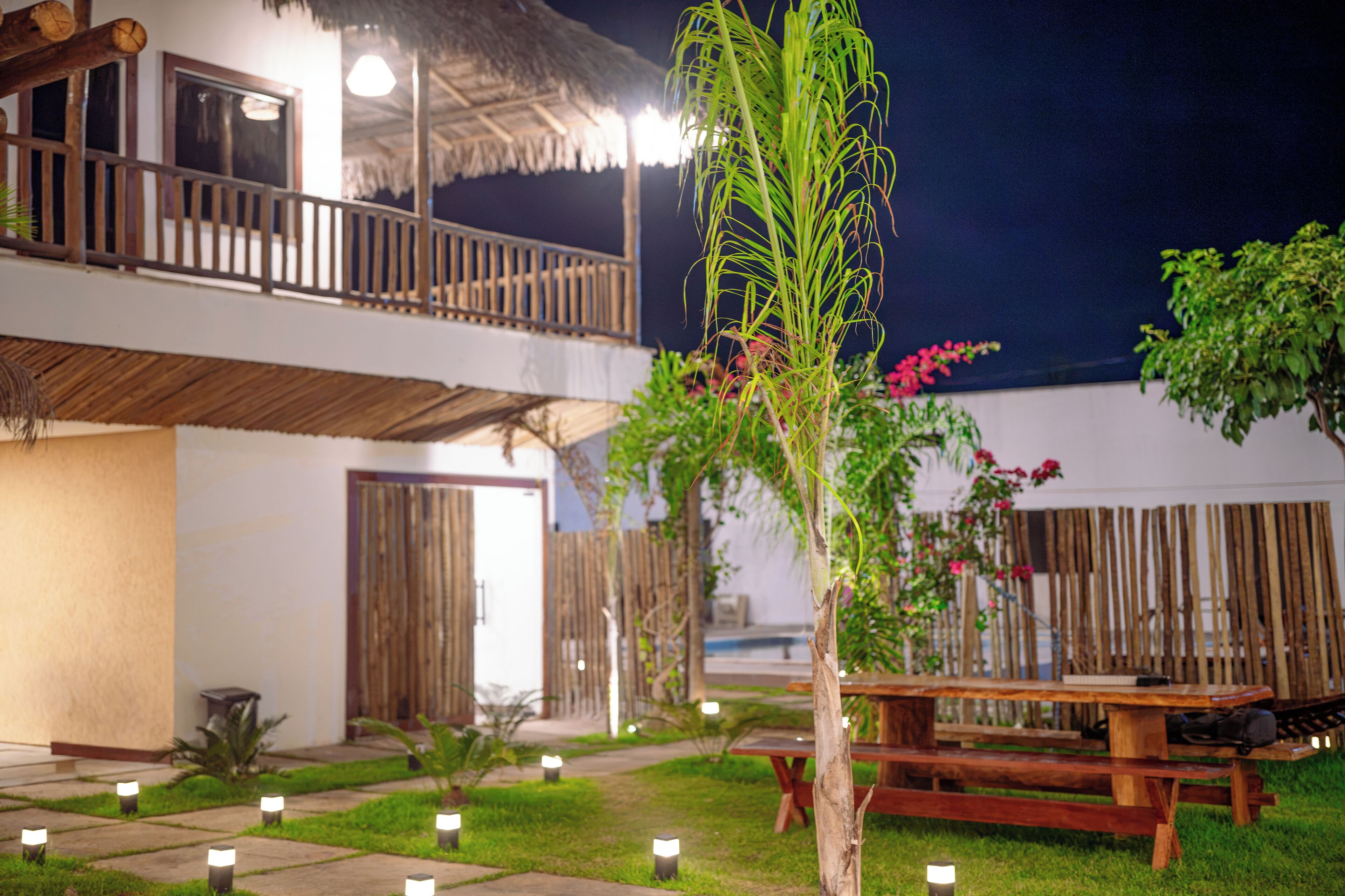 A well-lit outdoor area with a wooden deck, a thatched-roof structure, and a dining table.