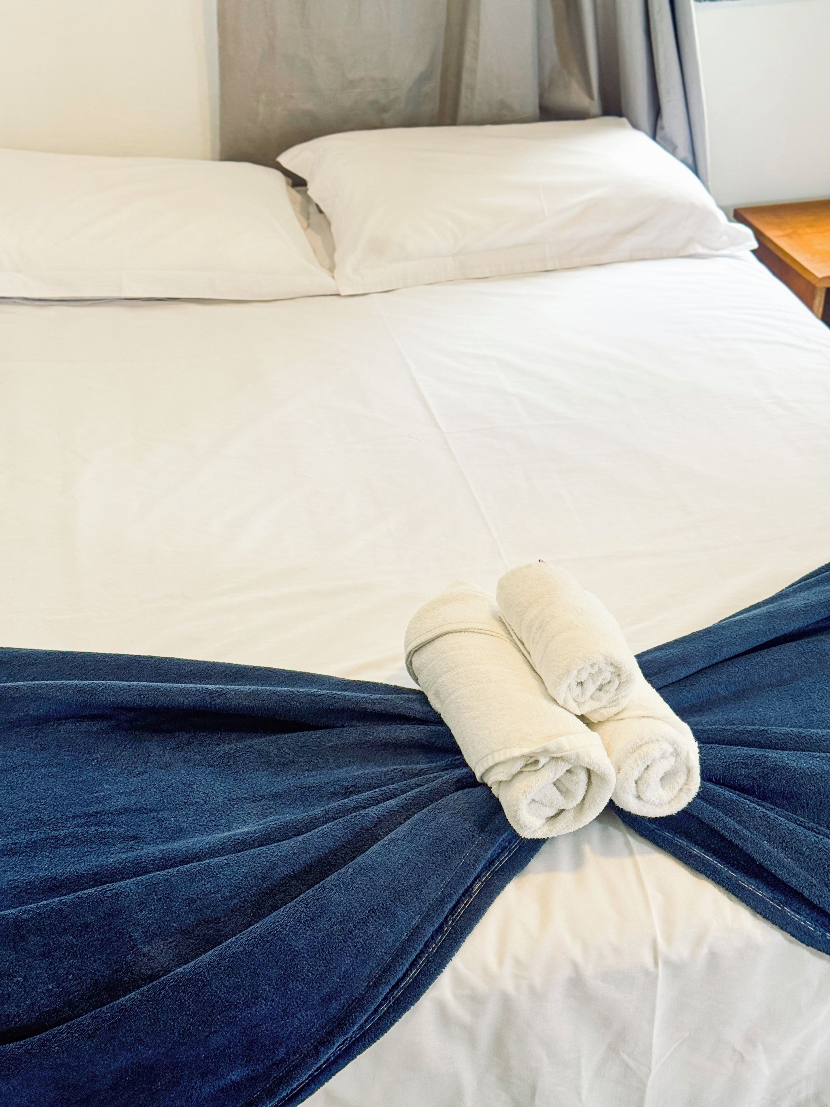 Una cama bien hecha con ropa blanca y una manta azul, con dos toallas enrolladas sobre la cama.