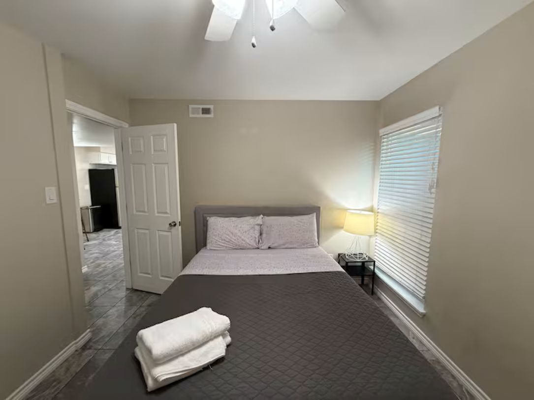A bedroom with a bed, a nightstand, a lamp, a window with blinds, and a door leading to another room.