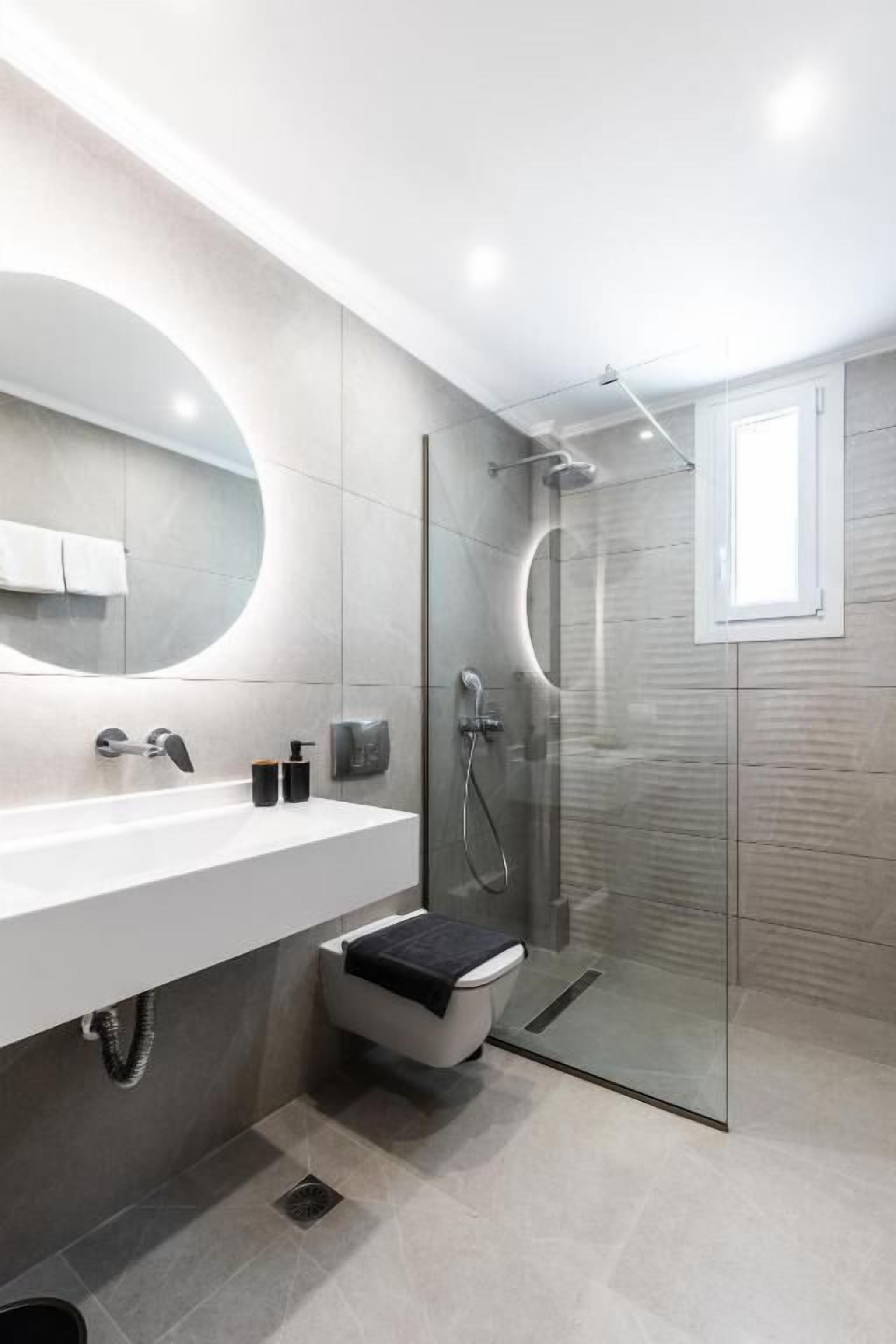 A modern bathroom with a large mirror, a white sink, a glass shower enclosure, and a small window.