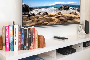A flat-screen TV mounted on a white entertainment center with a beach scene, surrounded by books and a remote control. - Stunning 1-bedroom condo in charming Kihei with AC Luana Kai C209 (Kihei)