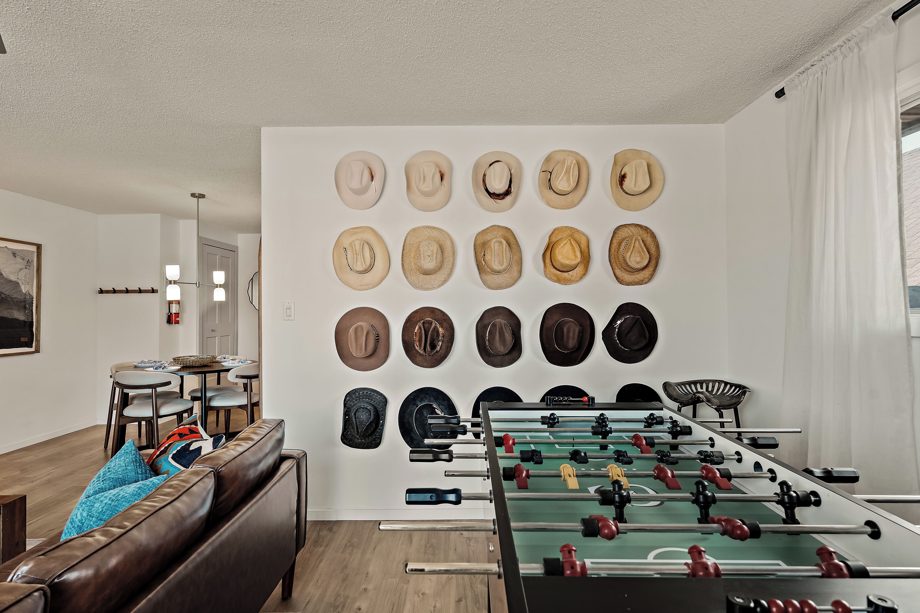 A room with a foosball table, a sofa with cushions, a dining table with chairs, and a wall with various hats.