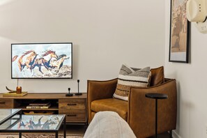 Ein Wohnzimmer mit einem braunen Ledersofa, einem Couchtisch, einem Fernseher, der ein Pferdegemälde zeigt, und einem Bücherregal mit Büchern.