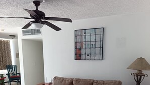 Een woonkamer met een beige bank, een zwart voetenbankje, een plafondventilator, een ingelijst kunstwerk en een lamp.