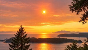 A sunset over a body of water with a forested island in the distance. - Sweeping Pacific Views & Unforgettable Sunsets · Your Island Escape, 20 Min Away (Bowen Island)