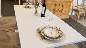 A dining area with a white table set for two, a cabinet with glass doors, and a bed in the background. - Sweeping Pacific Views & Unforgettable Sunsets · Your Island Escape, 20 Min Away (Bowen Island)