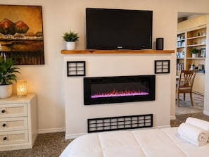 A bedroom with a fireplace, a television above it, a bed, a nightstand with a plant, and a bookshelf.